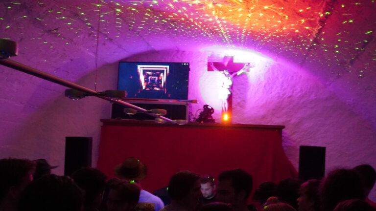 Junge Menschen feiern in einem bunt beleuchteten Gewölbekeller mit DJ-Pult und Kreuz an der Wand.