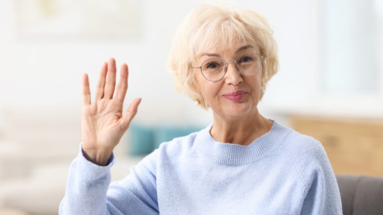 Eine alte Dame mit kurzen weißen Haaren winkt mit ihrer rechten Hand.