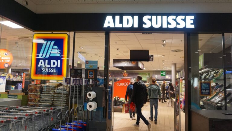 Abgebildet ist der Eingangsbereich einer Schweizer Aldi-Supermarktfiliale. Neben Einkaufswägen und eintretenden Kunden, sticht vor allem der Schriftzug „ALDI SUISSE“ in leuchtenden Lettern hervor.