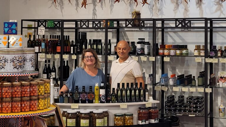 Hinter einer Ladentheke steht ein Ehepaar mittleren Alters umringt von Flaschen und Einmachgläsern.