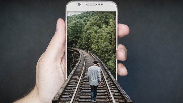 Auf einem Foto führen Bahngleise direkt in ein liegendes Smartphone, das im Vordergrund liegt. Ein Mann geht auf den Gleisen, die scheinbar in das Handy hineinfließen, wodurch das Bild symbolisch den Sog der digitalen Welt und den Druck durch Social Media auf den Einzelnen darstellt.