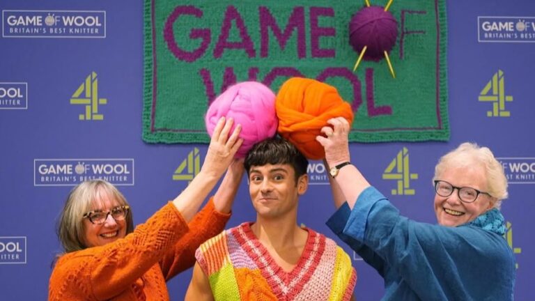 Zwei ältere Damen halten Wollknäuel über den Kopf eines jungen Mannes. Alle tragen gestrickte Pullover oder Cardigans.