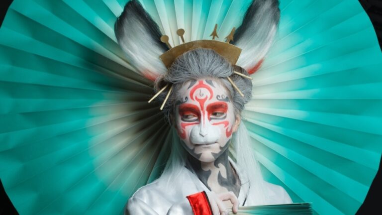 Eine Person in aufwendigem Cosplay trägt weiß-rote Gesichtsbemalung, tierähnliche Ohren und hält einen aufgefächerten, türkisfarbenen Fächer vor einem kreisförmigen Hintergrund.