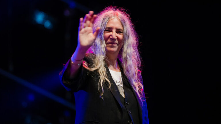 Die grauhaarige Patti Smith steht im lila-blauen Scheinwerferlicht und trägt einen schwarzen Anzug mit weißem Hemd darunter. Sie hebt grüßend ihre rechte Hand und schmunzelt zutraulich.