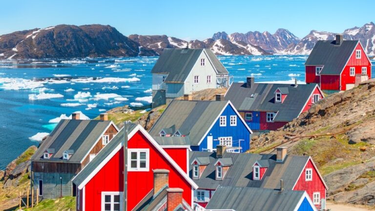 Rote, blaue und weiße Häuser an der Küste Grönlands. Im Hintergrund Meer mit Eisschollen und Berge.