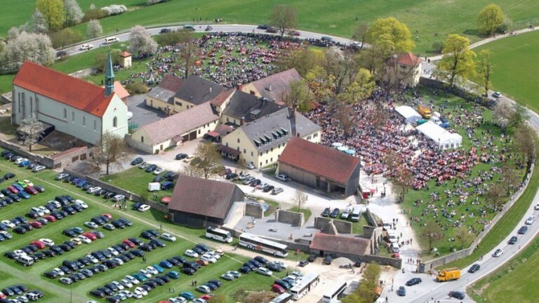 Aus der Vogelperspektive ist eine Kirche, mehrere Gebäude, geparkte Autos und ein ummauerter Hof mit hunderten Menschen darin zu sehen. Ringsum sind grüne Wiesen.