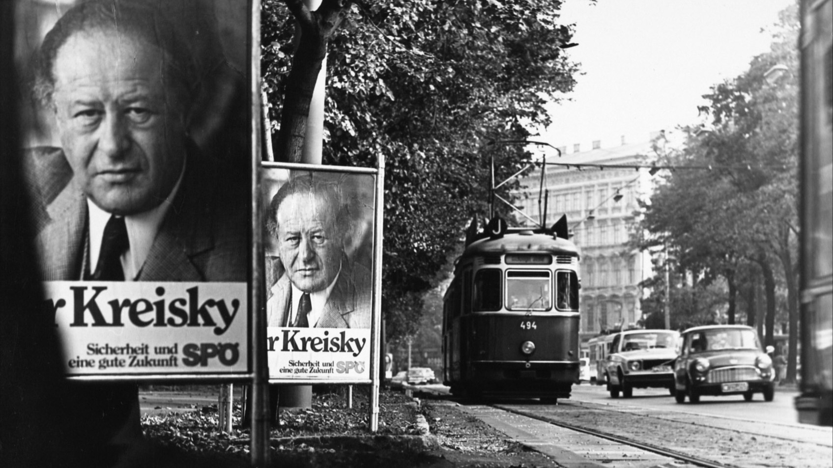 Schwarz-Weiß-Aufnahme einer Wiener Straßenbahn die an einer Reihe von Wahlplakaten mit dem Motiv Kruno Breiskys vorbeifährt.