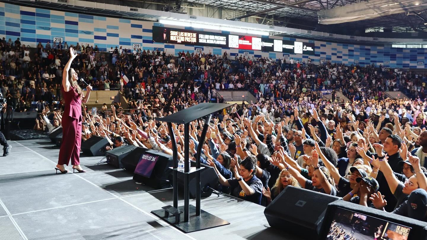 Kathryn Krick steht auf der Bühne einer Veranstaltungshalle und begrüßt hunderte Zuschauer.