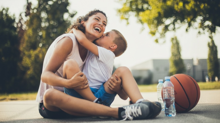 Eine lachende Mutter sitzt im Freien auf dem Boden und umarmt ihren Sohn herzlich. Neben ihnen liegen ein Basketball und Wasserflaschen auf dem Asphalt.
