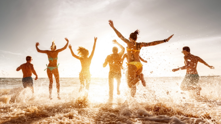 Das Bild zeigt mehrere Menschen in Badehose und Bikini, die am Strand der untergehenden Sonne entgegen laufen.