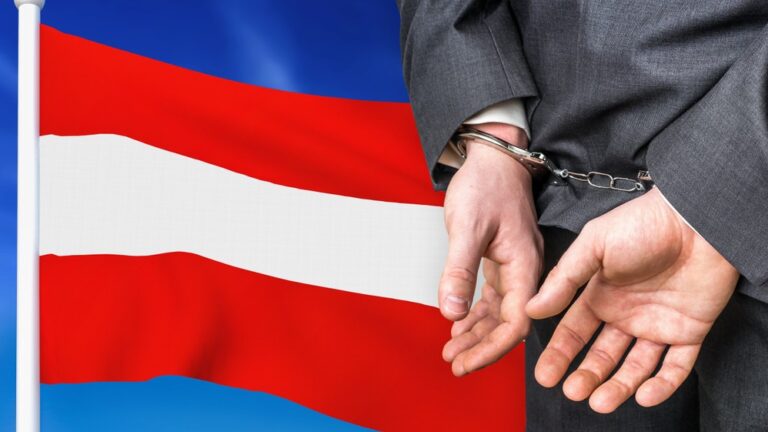 Das Bild zeigt auf der linken Seite die Rot-Weiß-Rote-Flagge Österreichs. Rechts im Bild sind zwei mit Handschellen gefesselte Hände (vermutlich eines Mannes) auf Gesäßhöhe. Man sieht, dass diese Person einen Anzug trägt und der Kamera den Rücken zugedreht hat.