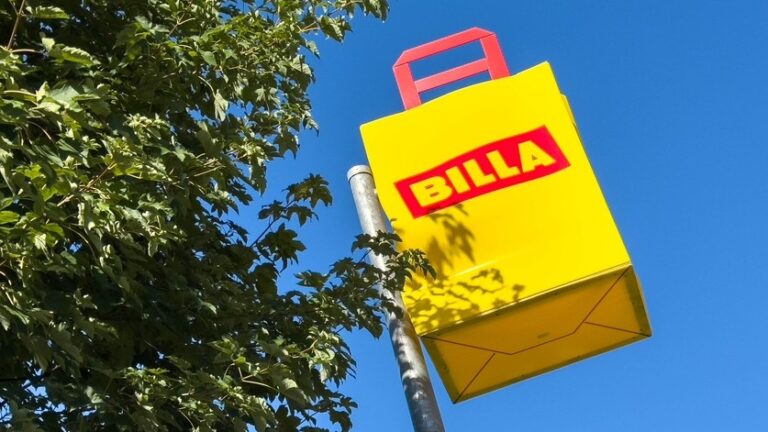 Gelbes Billa-Schild in Form einer Einkaufstasche an einem Metallpfosten vor blauem Himmel und Baumzweigen.