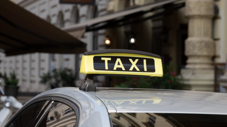 Dach eines Checker-Taxis (weiß) mit leuchtendem „Taxi“-Schild vor verschwommenem Hintergrund in Wiener Innenstadt