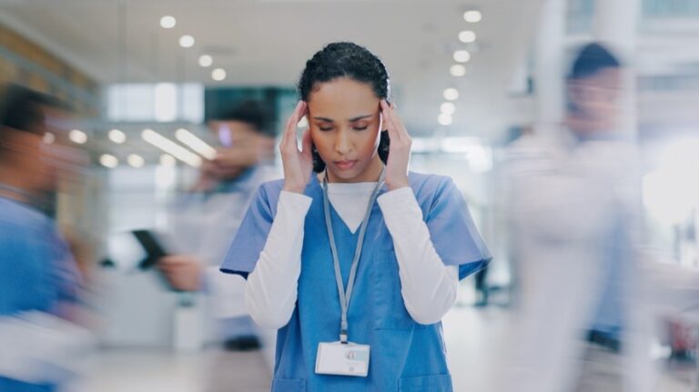 Eine Krankenpflegerin steht inmitten von Menschen, die so schnell an ihr vorbeilaufen, dass sie nur verschwommen erkennbar sind. Sie hält sich den Kopf, wirkt überfordert.