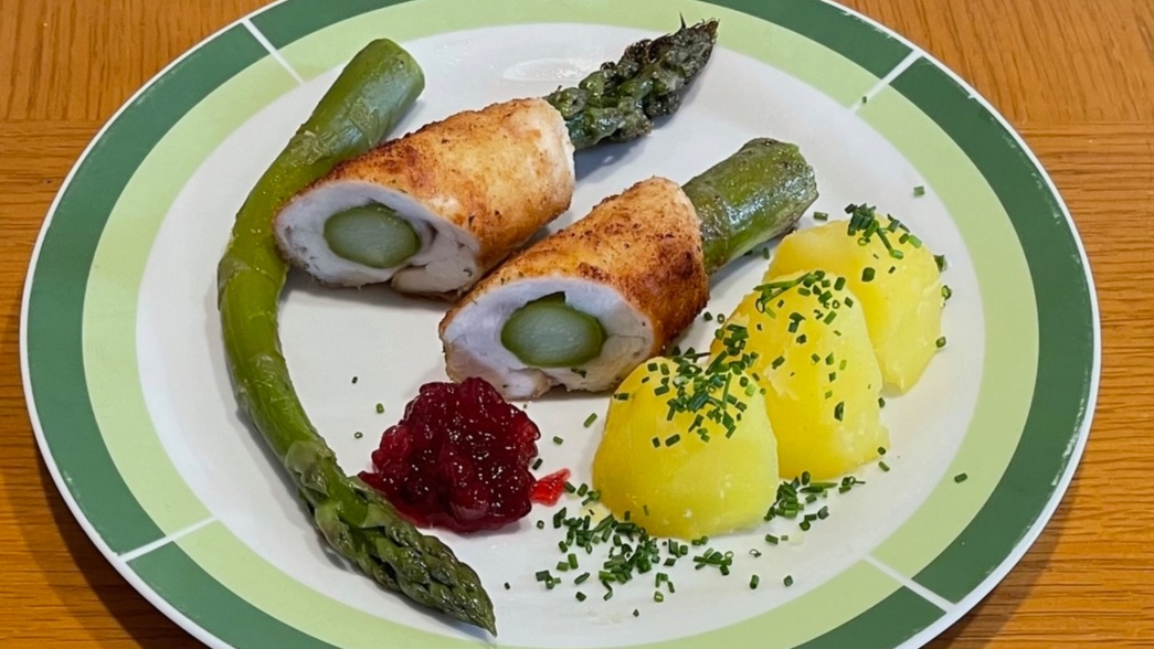 Spargel in paniertes Kaninchenfleisch eingerollt und zusammen mit Kartoffeln und Preiselbeeren auf einem Teller angerichtet.