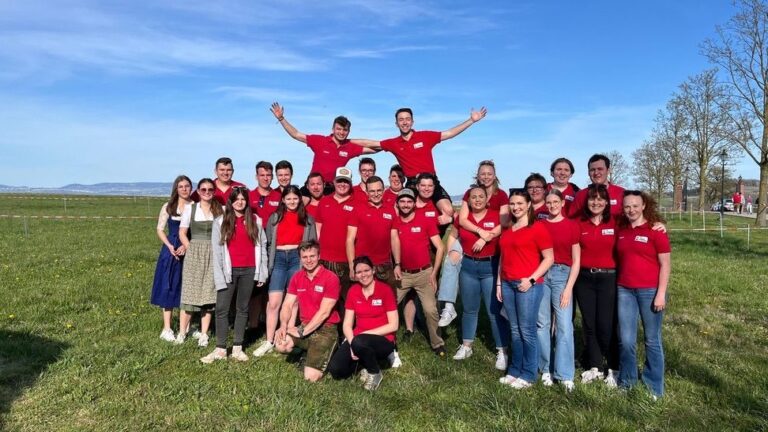 Eine Gruppe von Landjugend-Mitgliedern posiert lachend auf einer Wiese. Die Mehrheit trägt rote Shirts, zwei Personen tragen ein Dirndl. In der hinteren Reihe strecken zwei Personen die Arme in die Luft.