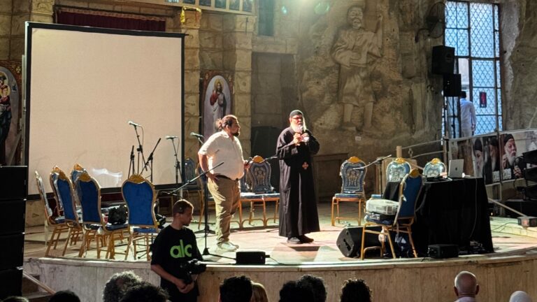 Jugendbischof Anba Moussa steht gemeinsam mit einem Dolmetscher auf einer Bühne in der Höhlenkirche von Manschiyyet Nasser in Kairo und spricht vor Publikum. Im Hintergrund sind die steinerne Kirchenwand, religiöse Darstellungen sowie Lichttechnik der Veranstaltung zu sehen.