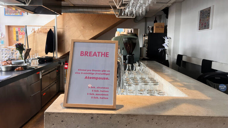 Blick auf die Theke des Cafés „Offline Oida“. Auf der Theke steht ein Schild mit der Aufschrift „Breathe“ und einer Anleitung zu einer Atemübung